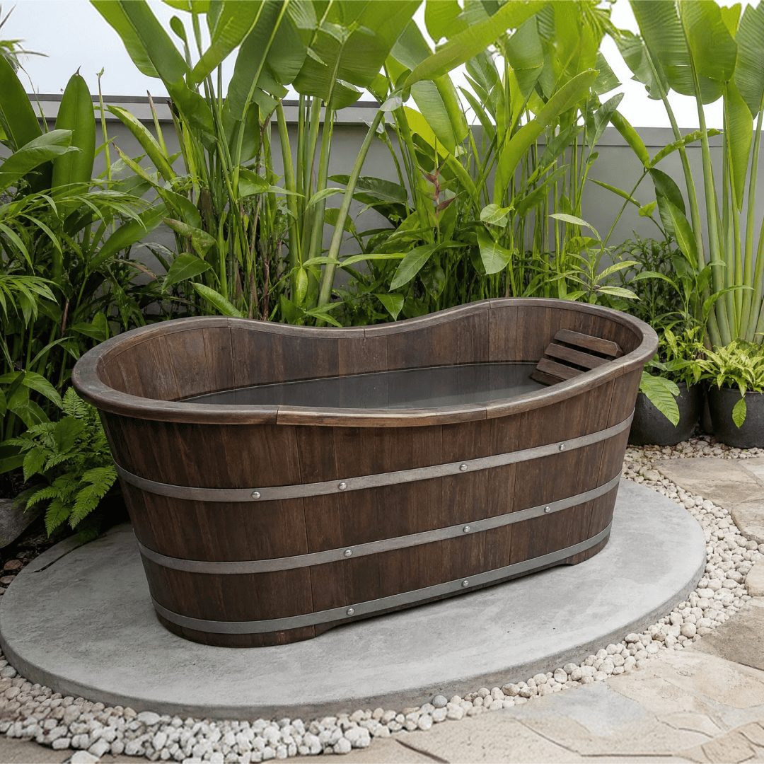 Dark timber ice bath filled with water on a pebble-edged concrete pad, framed by banana palms and heliconia