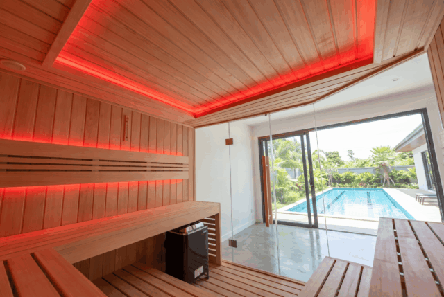 Sauna interior with red ceiling lighting, tiered timber benches and stone heater, pool visible through the glass door