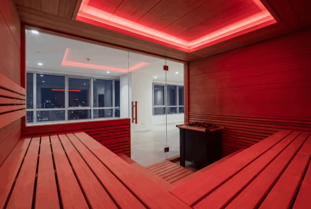 Sauna interior lit red throughout, slatted timber floor and benches, glass door looking out to daylight